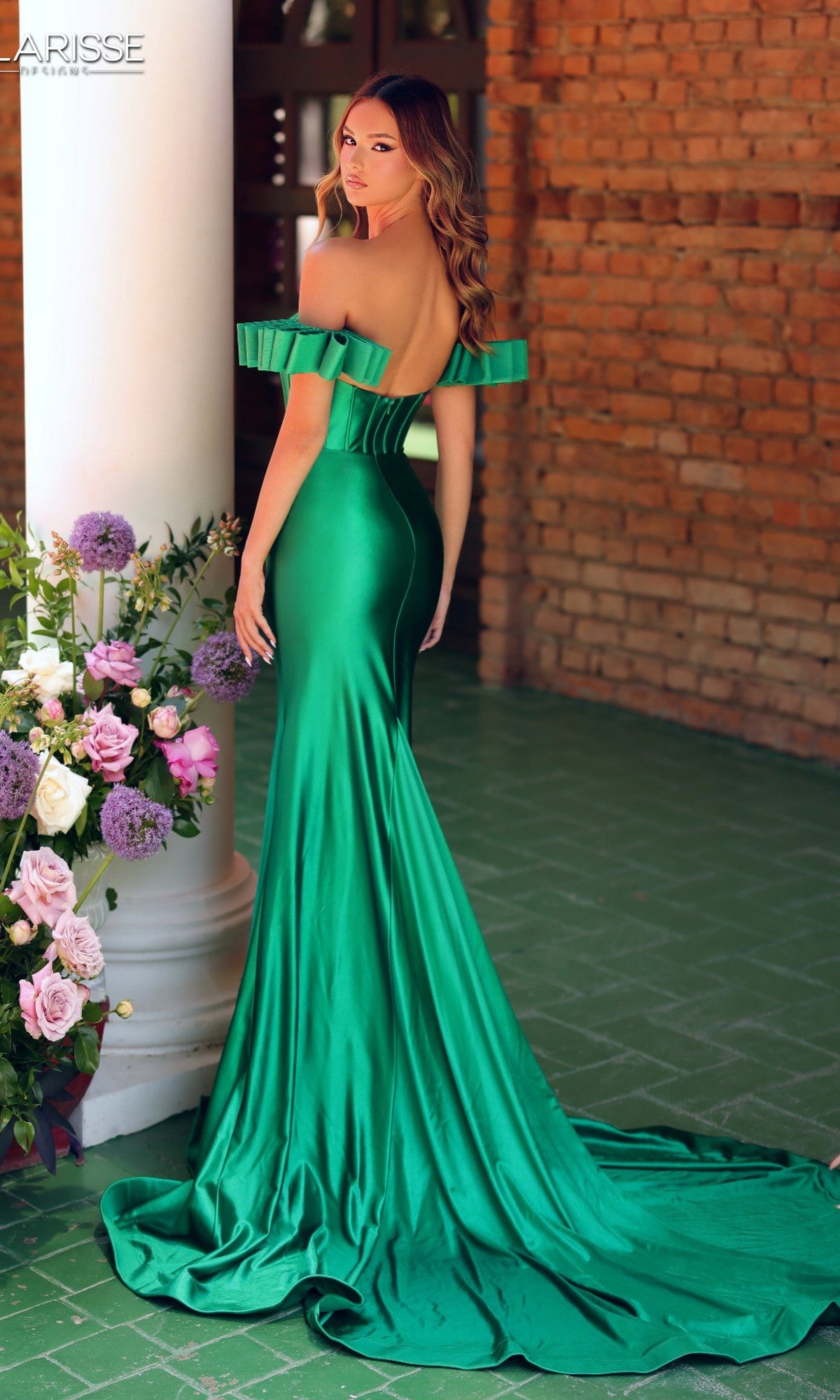 A woman in the Clarisse Designs 811160 long green satin prom dress stands sideways by a white column and floral arrangement against a brick wall.
